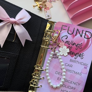 May include: A black textured binder with a light pink ribbon, featuring a pearl beaded charm with a heart and flower accents. A savings challenge insert lists categories like birthday, car, and vacation. A separate travel-themed gold keychain is nearby.