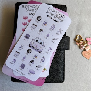 May include: A white Car Fund Savings Challenge tracker card featuring purple car-themed icons and dollar amounts, layered over a black binder and a Birthday Savings Challenge card. A gold-toned keychain with a heart charm labelled LOVE sits nearby.