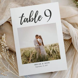 May include: A white wedding table number card with the text "table 9" in a handwritten style. The card features a photo of a couple embracing in a field. The names "ABIGAIL & ADAM" and the date "September 10, 2027" are printed below the photo.