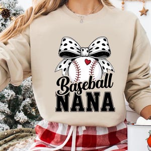 May include: Beige sweatshirt with a baseball graphic. The graphic features a baseball with a red heart, a black and white polka dot bow, and the words "Baseball NANA" in black script and block letters.