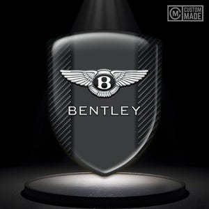 Puede incluir: Un emblema en forma de escudo con el logotipo de Bentley, con un emblema alado plateado sobre el nombre de la marca. El fondo tiene una textura de fibra de carbono. El emblema se muestra en un escenario oscuro con un foco.