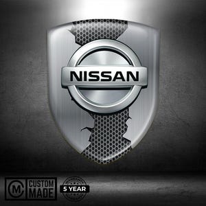 Op de afbeelding: Een zilveren Nissan-embleem, schildvormig, met de merknaam prominent weergegeven. Het embleem heeft een getextureerde achtergrond en een geperforeerd gedeelte. De woorden "Custom Made" en "5 Year Warranty" zijn ook zichtbaar.