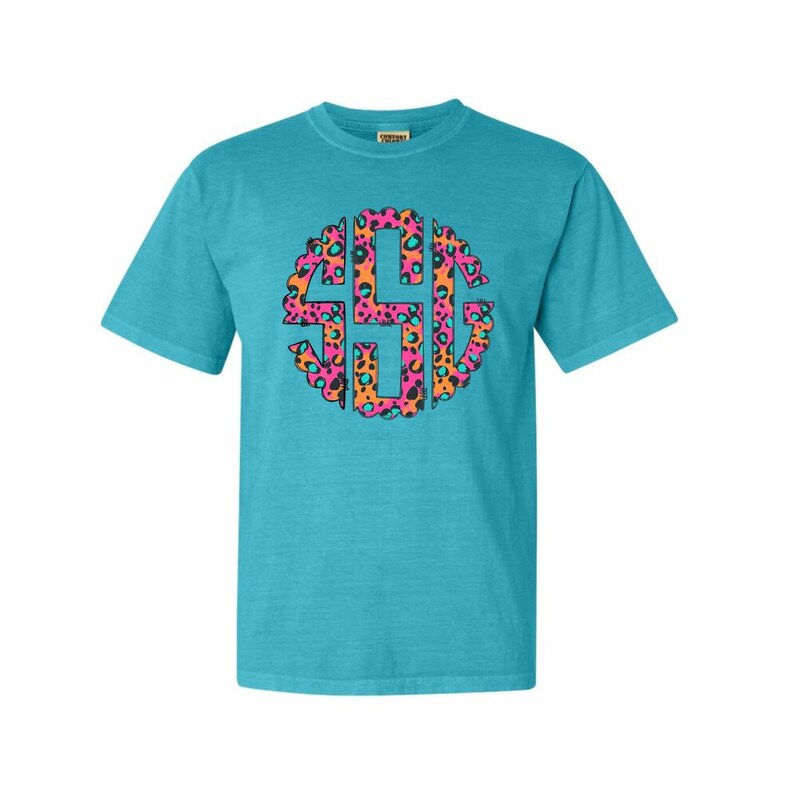 Op de afbeelding: Een turquoise t-shirt met korte mouwen met een kleurrijk monogram met luipaardprint. Het monogram is cirkelvormig, met roze, oranje en turquoise accenten. Het shirt is gemaakt van een zacht, comfortabel materiaal.