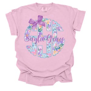 Puede incluir: Camiseta rosa claro con un diseño de monograma floral y un lazo morado. El monograma incluye el nombre "Saylee Grace" en cursiva. El diseño floral incorpora flores azules, moradas y amarillas.