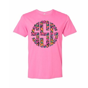 Op de afbeelding: Een roze t-shirt met een cirkelvormig luipaardprintontwerp met de letters "SHG". De luipaardprint bevat oranje, roze en turquoise tinten. Het t-shirt zelf is effen roze.