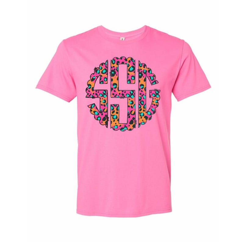 Op de afbeelding: Een roze t-shirt met een cirkelvormig luipaardprintontwerp met de letters "SHG". De luipaardprint bevat oranje, roze en turquoise tinten. Het t-shirt zelf is effen roze.