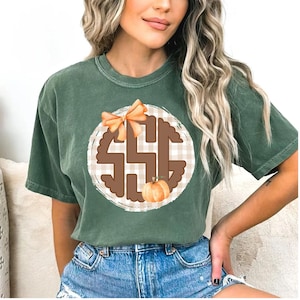May include: Green t-shirt featuring a circular design with the letters "SSG" in brown, a plaid background, a pumpkin, and an orange bow. The shirt is a solid green color.
