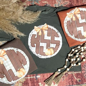 Könnte beinhalten: Drei T-Shirts in Braun- und Grüntönen, jedes mit einem kreisförmigen Design mit Monogramm, Schleife und Kürbis. Eine Perlenkette und getrocknetes Gras sind ebenfalls sichtbar. Die Shirts sind wahrscheinlich für den Herbst.