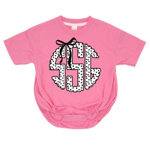 May include: Pink baby bodysuit with a white and black polka dot monogram design. The monogram is in a scalloped circle shape and features a black bow at the top. The bodysuit has short sleeves and snap closures.