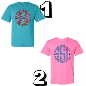 Op de afbeelding: Twee T-shirts met korte mouwen. E&eacute;n is turquoise met een cirkelvormig monogram in oranje, roze en zwart luipaardprint. De andere is roze met een cirkelvormig monogram in blauwe, roze en gele bloemenprint.