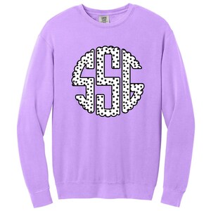 May include: A light purple sweatshirt with a large, circular monogram design. The monogram features the letters "SSG" in a white, dotted pattern, outlined with a scalloped edge. The sweatshirt has long sleeves and a crew neck.