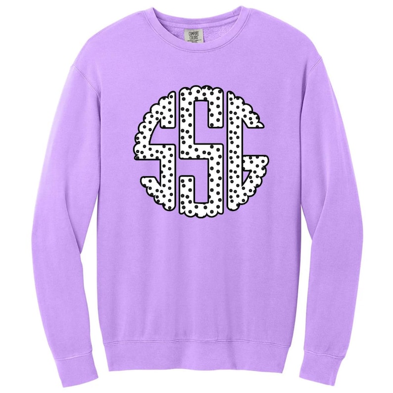 May include: A light purple sweatshirt with a large, circular monogram design. The monogram features the letters "SSG" in a white, dotted pattern, outlined with a scalloped edge. The sweatshirt has long sleeves and a crew neck.