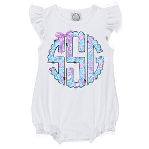 May include: White baby romper with ruffled sleeves. The front features a monogram design in a scalloped circle, with a light blue background and pink roses and hearts. A purple bow is at the top of the monogram.