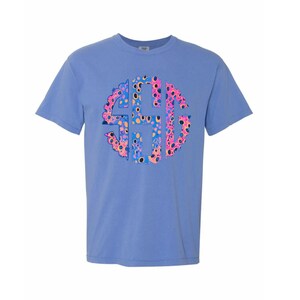 Op de afbeelding: Een lichtblauw t-shirt met een kleurrijk monogramontwerp. Het monogram is een cirkelvormig ontwerp met de letters "SGH" in een levendig patroon van roze, blauwe, gele en zwarte stippen. Het shirt heeft korte mouwen en een ronde hals.