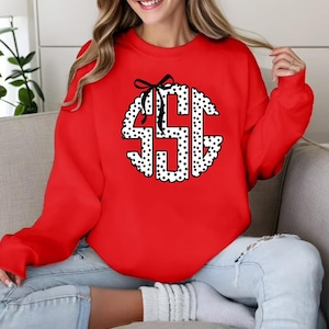 May include: A red sweatshirt featuring a white and black polka-dot monogram design. The monogram is in a circular shape with a black bow at the top. The sweatshirt is a solid red color.