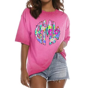 May include: A hot pink t-shirt with a large, colorful floral monogram design. The monogram features the letters "GML" in a circular pattern, with white daisy accents. The shirt has short sleeves and a relaxed fit.