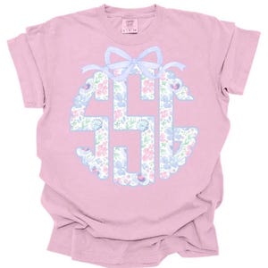 May include: A light pink t-shirt featuring a floral monogram design in blue, pink, and white. The monogram is encircled by a scalloped border and topped with a light blue bow. The shirt is made of a soft material.