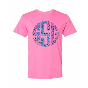 Op de afbeelding: Een roze t-shirt met een cirkelvormig monogramontwerp. Het monogram is gevuld met een kleurrijk patroon van blauwe, gele en roze stippen en vormen. Het shirt heeft een effen roze kleur.