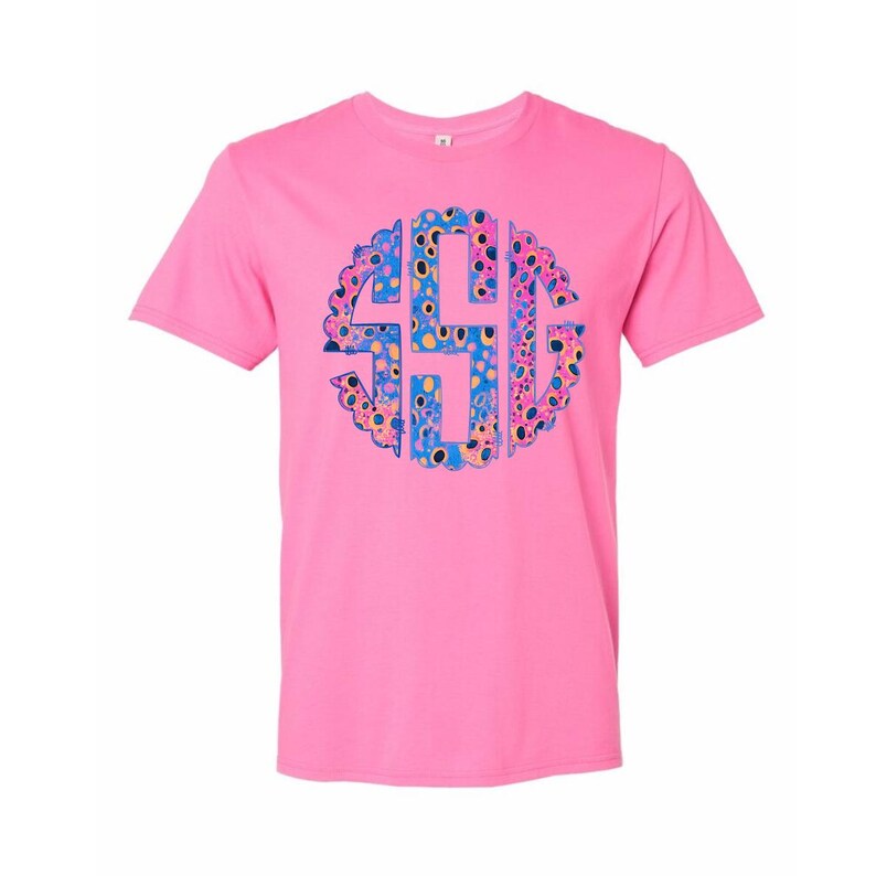 Op de afbeelding: Een roze t-shirt met een cirkelvormig monogramontwerp. Het monogram is gevuld met een kleurrijk patroon van blauwe, gele en roze stippen en vormen. Het shirt heeft een effen roze kleur.