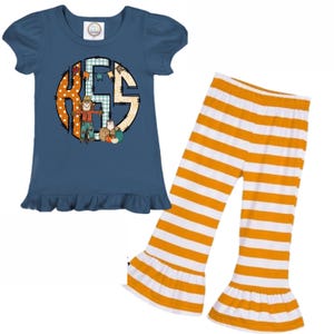 May include: A two-piece outfit featuring a navy blue short-sleeved top with a ruffle hem and a circular graphic with a scarecrow and pumpkins. The top has a monogram design. The outfit includes orange and white striped bell-bottom trousers.