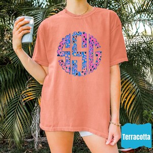 Op de afbeelding: Terracotta-kleurig T-shirt met een groot, cirkelvormig monogramontwerp in blauw en roze. Het monogram bestaat uit drie letters. Het shirt heeft korte mouwen en een relaxte pasvorm. Het woord "Terracotta" staat in een blauwe doos rechtsonder.