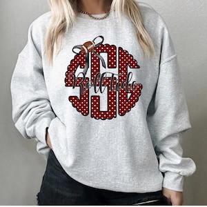 May include: A light gray sweatshirt featuring a red and white polka-dot monogram with the words "Roll Tide" in a script font. A football and bow graphic is at the top of the monogram. The sweatshirt is a crew neck style.