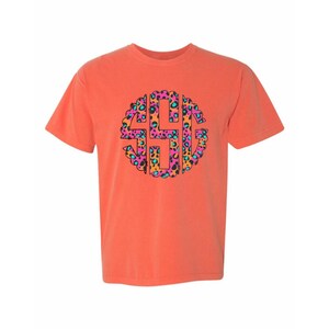 Op de afbeelding: Koraalrood T-shirt met een cirkelvormig monogramontwerp. Het monogram is gevuld met een luipaardprintpatroon in roze, oranje, teal en zwart. Het shirt heeft korte mouwen en een ronde hals.