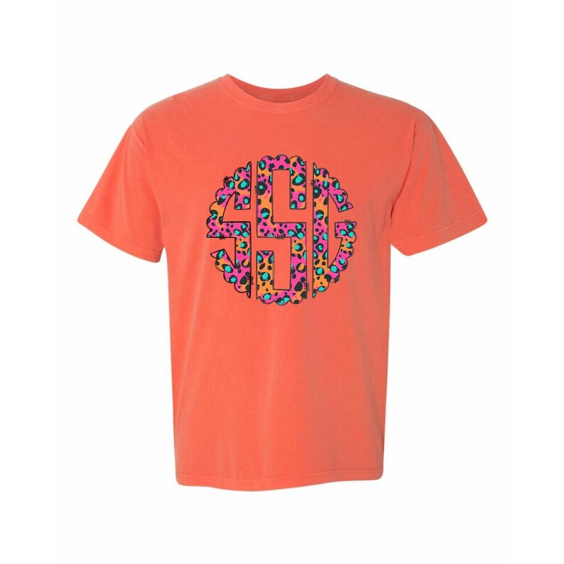 Op de afbeelding: Koraalrood T-shirt met een cirkelvormig monogramontwerp. Het monogram is gevuld met een luipaardprintpatroon in roze, oranje, teal en zwart. Het shirt heeft korte mouwen en een ronde hals.