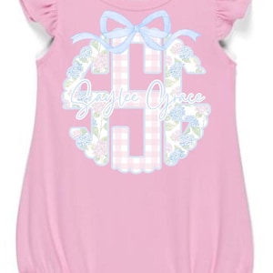 Könnte beinhalten: Rosa Baby-Strampler mit Rüschenärmeln. Verfügt über eine hellblaue Schleife und ein Monogramm mit dem Namen "Saylee Grace" in floralem Design. Der Strampler hat eine Ballon-Silhouette.