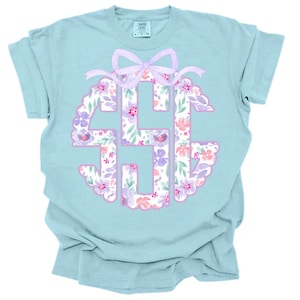 May include: A pale blue t-shirt with a floral monogram design. The monogram is in a scalloped circle with a lilac bow at the top. The monogram is filled with pink and purple flowers and green leaves.