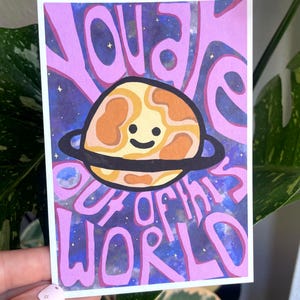 Puede incluir: Una impresión artística rectangular con las palabras "YOU ARE OUT OF THIS WORLD" en letras rosas. La impresión presenta un planeta sonriente con un anillo negro, sobre un fondo estrellado, morado y azul.