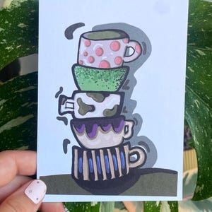 Könnte beinhalten: Eine skurrile Illustration eines Stapels von vier bunten Teetassen. Die oberste Tasse ist rosa mit roten Punkten, gefolgt von einer grünen Tasse, einer weißen Tasse mit braunen Flecken und einer gestreiften Tasse mit blauen und violetten Akzenten. Das Kunstwerk befindet sich auf einer weißen Karte.