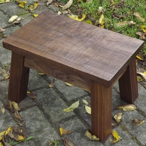 May include: A dark brown wooden stool with a rectangular top and four legs. The stool is sitting on a brick patio with fallen leaves.