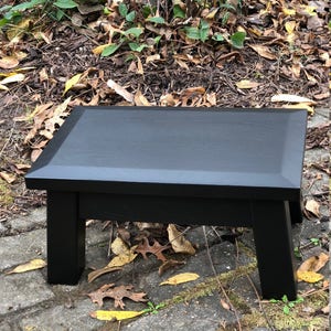 May include: A black wooden step stool with a rectangular top and four legs. The stool is sitting on a brick patio with leaves scattered around it.