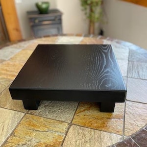 May include: A square, black-stained wooden riser with a visible wood grain texture. The platform is elevated by four short, block-style legs, providing a flat, raised surface for display purposes. The finish is smooth with a matte sheen.