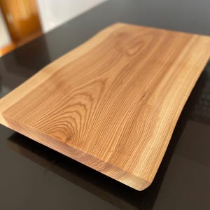May include: A large, natural wood cutting board with a live edge and a smooth, polished surface. The board has a light brown color and a natural wood grain pattern.