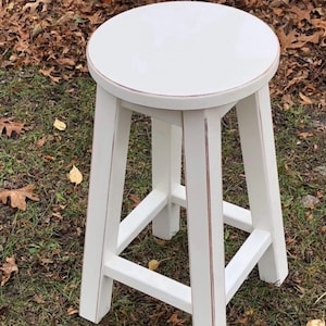 May include: White wooden stool with a round seat and a distressed finish. The stool has a simple design with four legs and a single stretcher.