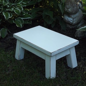 May include: White wooden step stool with a distressed finish. The stool has a square top and four legs.