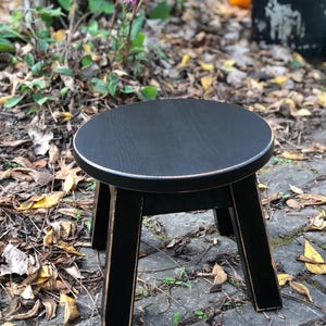 May include: A black wooden stool with a round top and three legs. The stool has a distressed finish, giving it a vintage look.