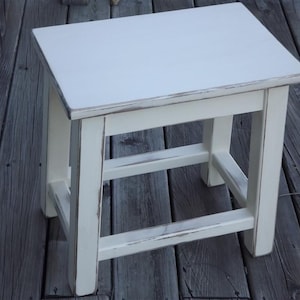 May include: White wooden stool with a distressed finish. The stool has a simple design with four legs and a rectangular top.