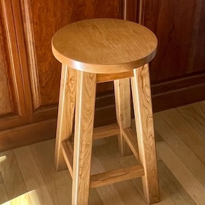 May include: A wooden stool with a round seat and a simple, sturdy design. The stool is made of light-colored wood with a natural finish.