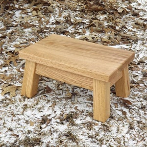 May include: A small wooden stool with a natural wood finish. The stool has four legs and a rectangular top.