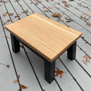 May include: A small wooden stool with a natural wood top and black painted legs. The stool is sitting on a wooden deck.