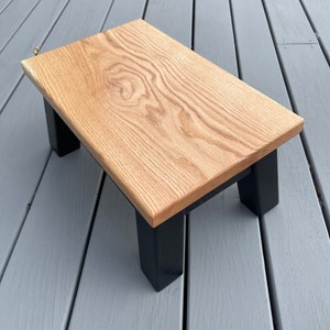 May include: A small wooden stool with a natural wood top and black painted legs. The stool is sitting on a gray wooden deck.