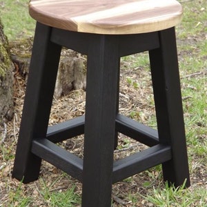 May include: A black wooden stool with a round, oval-shaped walnut wood top. The stool has a simple, sturdy design with a square base and four legs.