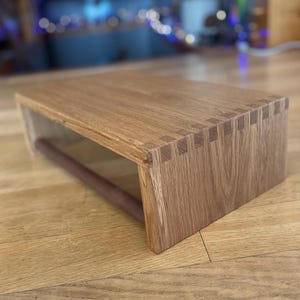 May include: A rectangular wooden paper towel holder. The holder is made of light brown wood with visible grain and features a dark brown dowel for holding the paper towels. The corners are joined with dovetail joints.