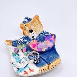 May include: A ceramic bear-shaped jewelry dish with a blue uniform design. The dish holds a variety of jewelry, including earrings with green beads and a colorful pendant. A silver ring and a blue bird illustration are also visible.