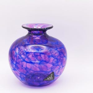 Op de afbeelding: Een ronde, handgemaakte glazen vaas met een brede body en een smalle hals. De vaas heeft een wervelend patroon van paarse en blauwe kleuren. De vaas heeft een zwarte rand en een label met de tekst "Kerry Glass Handmade in Ireland."