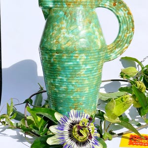 Könnte beinhalten: Eine grüne Vintage-Keramikkrug mit einem großen Griff. Der Krug hat eine strukturierte Oberfläche mit braunen Akzenten. Eine Passionsblume und Grünzeug befinden sich im Vordergrund. Der Krug ist wahrscheinlich für dekorative Zwecke.