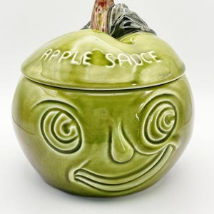 Może przedstawiać: Zielony ceramiczny słoik na mus jabłkowy z pokrywką. Słoik ma wzór twarzy ze spiralnymi oczami i uśmiechniętymi ustami. Na pokrywce widnieje napis "APPLE SAUCE". Na górze słoika znajduje się wzór łodygi i liścia.